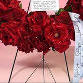 A circular floral wreath made of deep red roses with green leaves, displayed on a black metal stand against a solid pink background. A white handwritten card and a white ribbon with black script are attached to the wreath.
