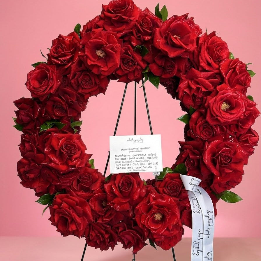 A circular floral wreath made of deep red roses with green leaves, displayed on a black metal stand against a solid pink background. A white handwritten card and a white ribbon with black script are attached to the wreath.