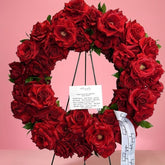 A circular floral wreath made of deep red roses with green leaves, displayed on a black metal stand against a solid pink background. A white handwritten card and a white ribbon with black script are attached to the wreath.