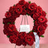 A circular floral wreath made of deep red roses with green leaves, displayed on a black metal stand against a solid pink background. A white handwritten card and a white ribbon with black script are attached to the wreath.