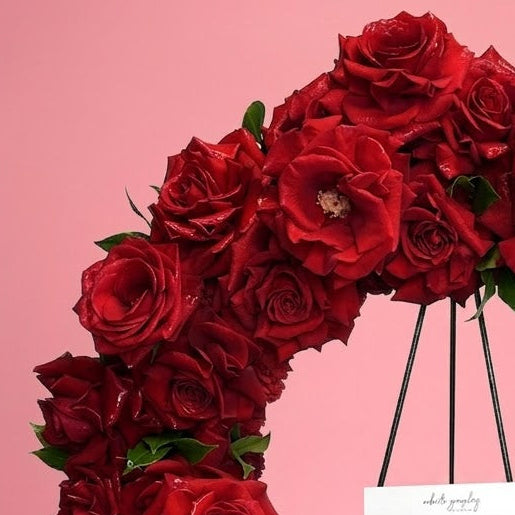 A circular floral wreath made of deep red roses with green leaves, displayed on a black metal stand against a solid pink background. A white handwritten card and a white ribbon with black script are attached to the wreath.
