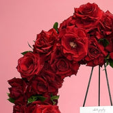 A circular floral wreath made of deep red roses with green leaves, displayed on a black metal stand against a solid pink background. A white handwritten card and a white ribbon with black script are attached to the wreath.