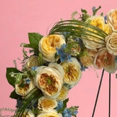 A circular floral wreath with yellow and cream roses, blue filler flowers, and green foliage, displayed on a black metal stand against a solid pink background.