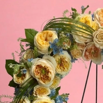 A circular floral wreath with yellow and cream roses, blue filler flowers, and green foliage, displayed on a black metal stand against a solid pink background.