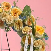 A circular floral wreath with yellow and cream roses, blue filler flowers, and green foliage, displayed on a black metal stand against a solid pink background.