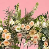 A large floral wreath on a black metal stand against a solid pink studio background. The wreath features white and peach roses, green foliage, and small white berries, with a small white card attached at the center.