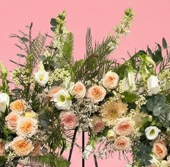 A large floral wreath on a black metal stand against a solid pink studio background. The wreath features white and peach roses, green foliage, and small white berries, with a small white card attached at the center.