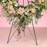 A large floral wreath on a black metal stand against a solid pink studio background. The wreath features white and peach roses, green foliage, and small white berries, with a small white card attached at the center.