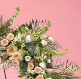 A large floral wreath on a black metal stand against a solid pink studio background. The wreath features white and peach roses, green foliage, and small white berries, with a small white card attached at the center.