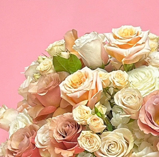 A large, lush floral arrangement featuring white, pink, and peach roses with hydrangea accents, presented on a silver pedestal stand against a solid pink background.