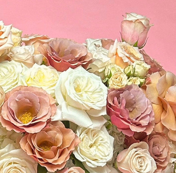 A large, lush floral arrangement featuring white, pink, and peach roses with hydrangea accents, presented on a silver pedestal stand against a solid pink background.