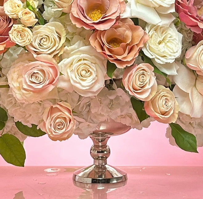 A large, lush floral arrangement featuring white, pink, and peach roses with hydrangea accents, presented on a silver pedestal stand against a solid pink background.