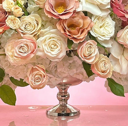 A large, lush floral arrangement featuring white, pink, and peach roses with hydrangea accents, presented on a silver pedestal stand against a solid pink background.